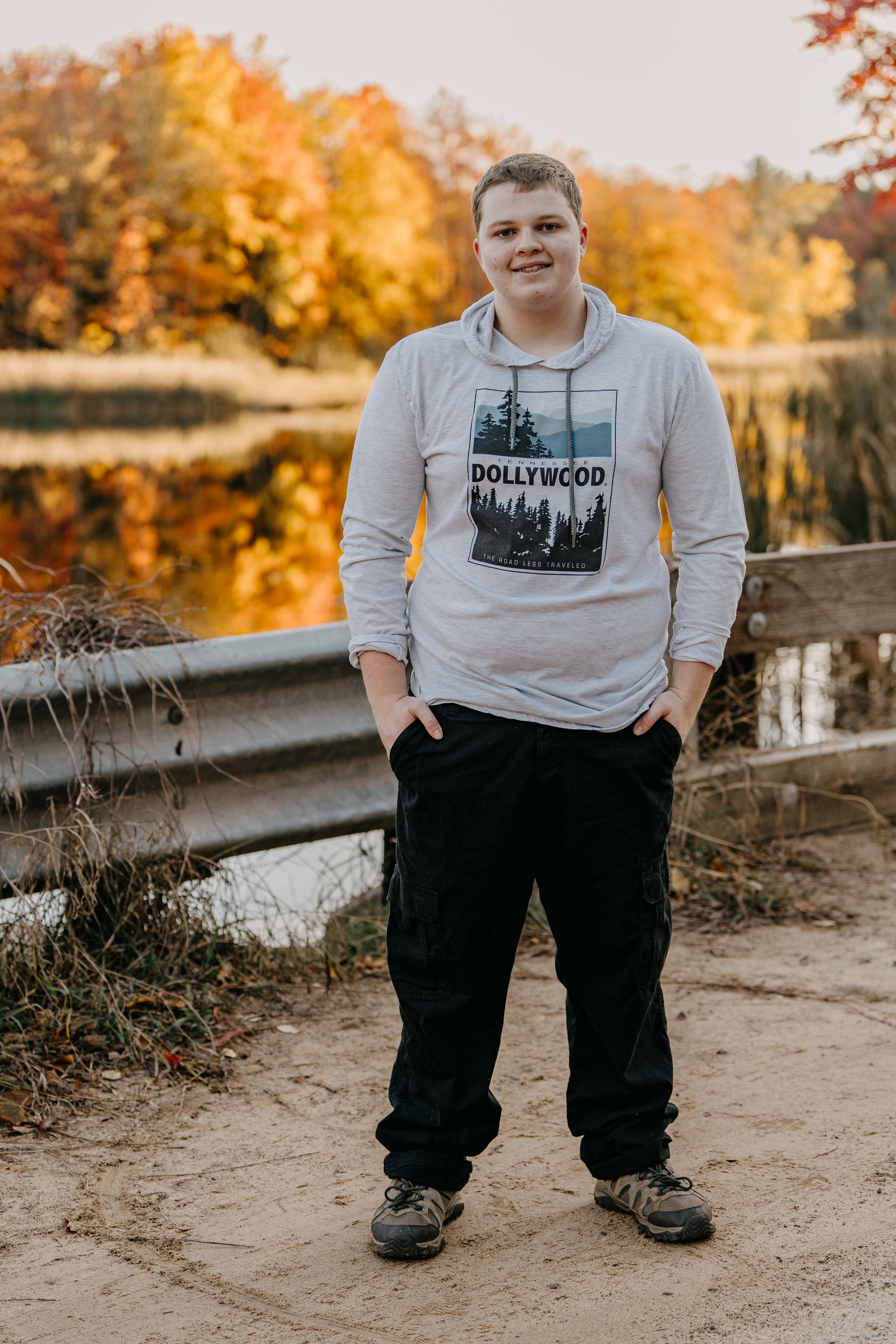 A person in a Hollywood sweatshirt stands on a path with vibrant autumn foliage reflecting in the lake behind.