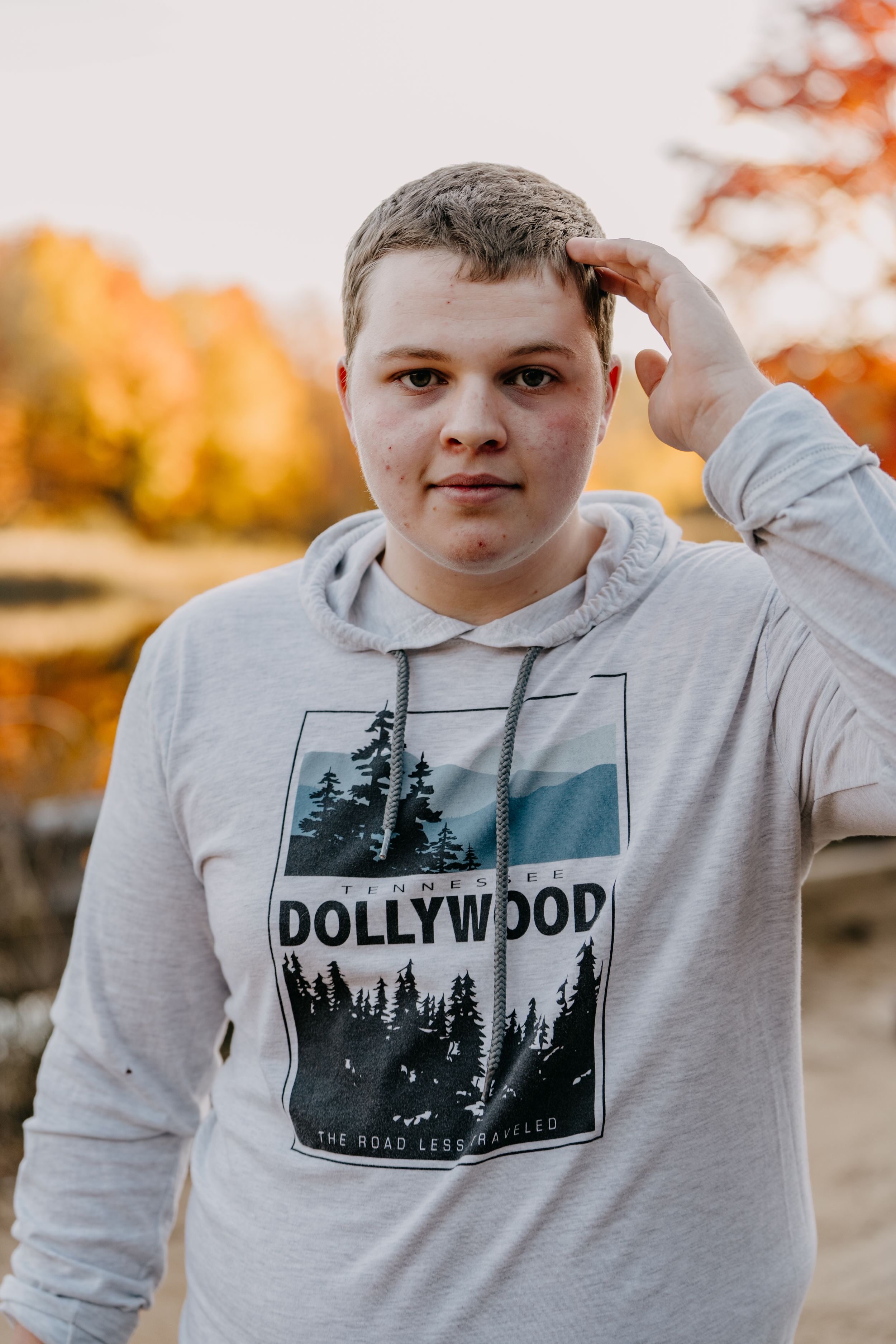 Person wearing Dollywood graphic hoodie poses outdoors with fall foliage in background.