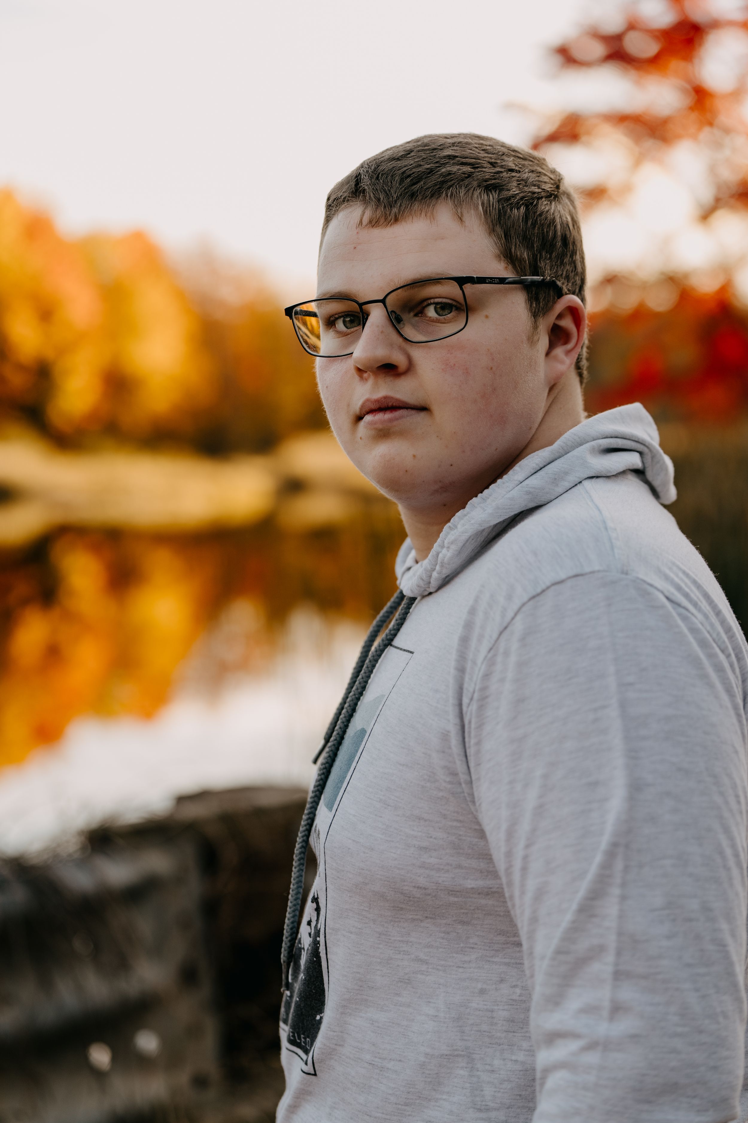 Portrait of a young person in gray hoodie and glasses against fall foliage and reflective water background.
