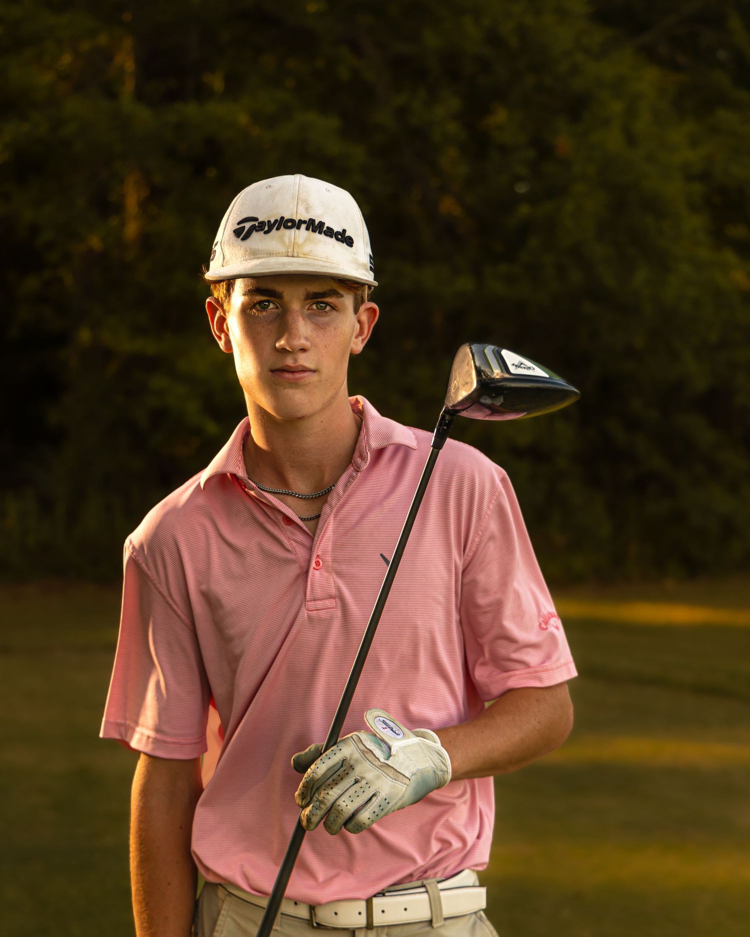A golfer in a pink polo shirt and khaki shorts swings a driver during golden hour on a golf course.