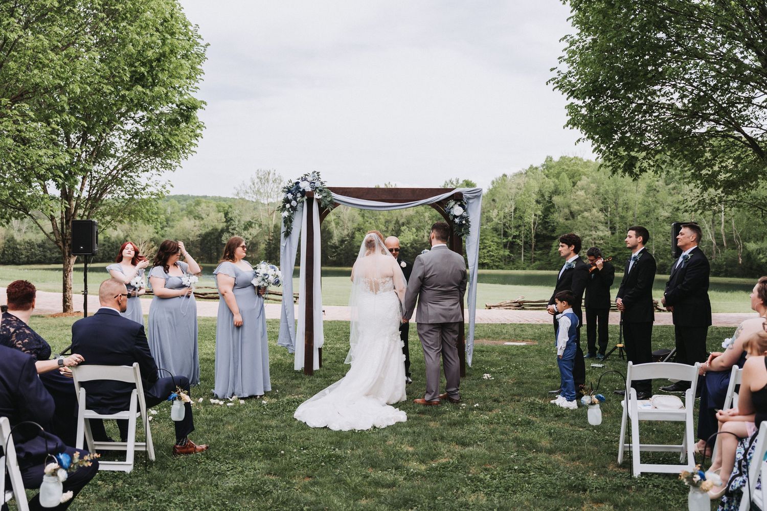 An outdoor wedding ceremony taking place on a grassy lawn with guests seated in white chairs and a decorative arch.