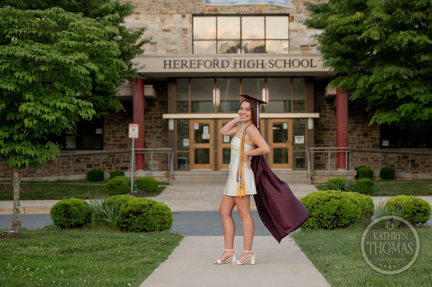 Graduate in a beige dress poses outside the stone facade of Hereford High School with graduation gown on arm.