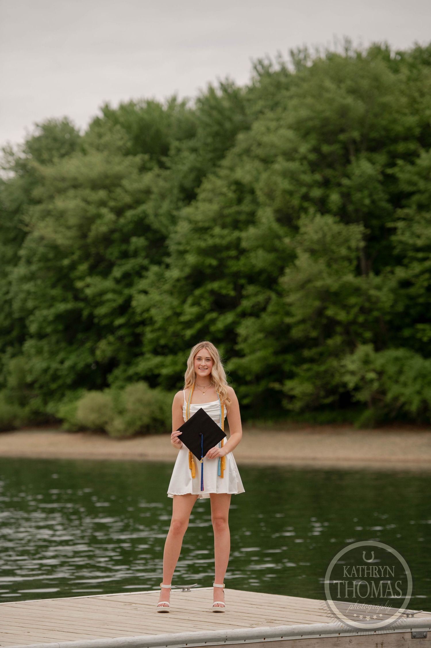 A graduate in a white dress stands on a wooden dock by a lake surrounded by lush green trees on a summer day.