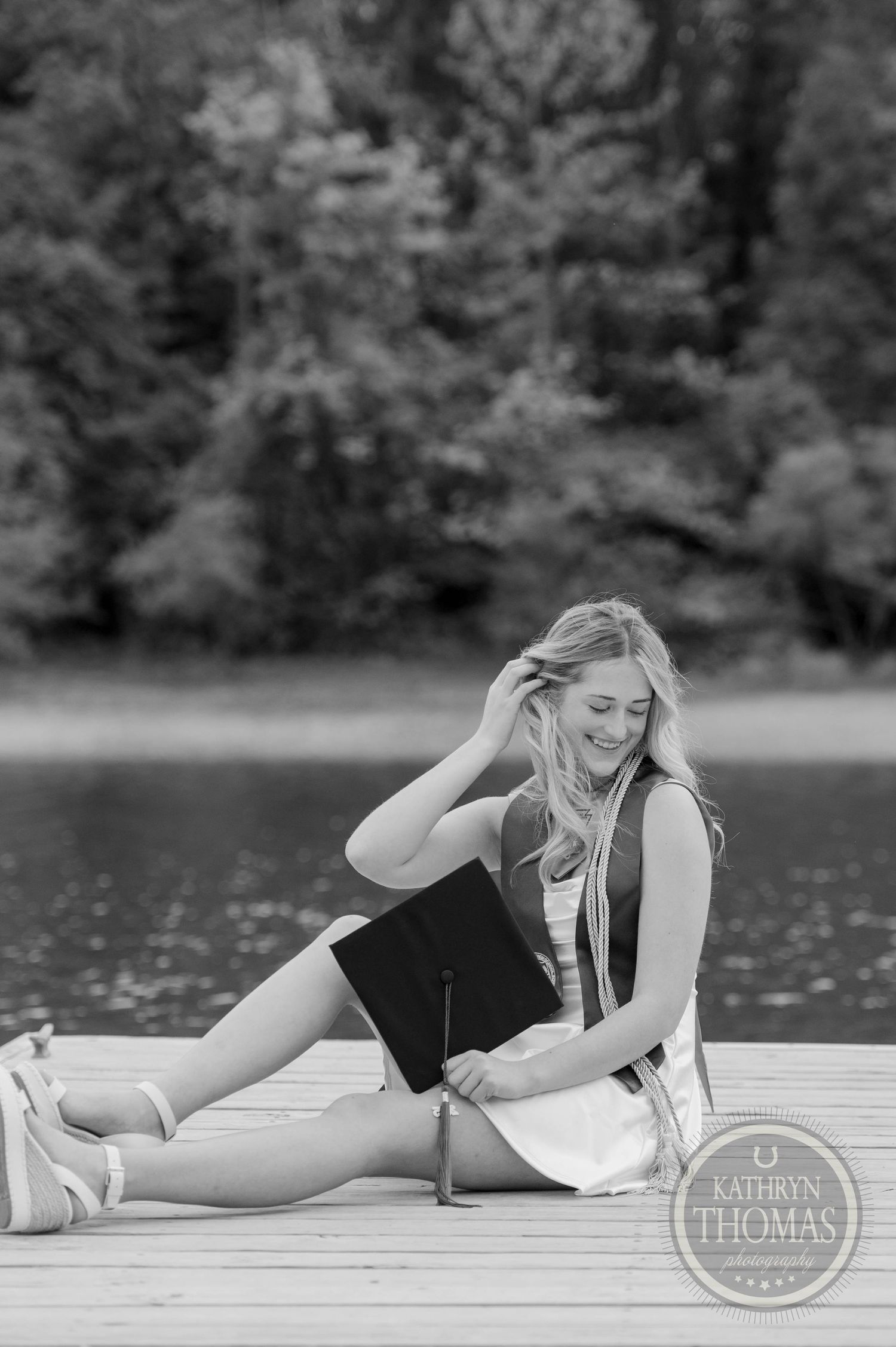 A joyful figure sits on a wooden dock by a peaceful lake, wearing casual summer attire in black and white photography.