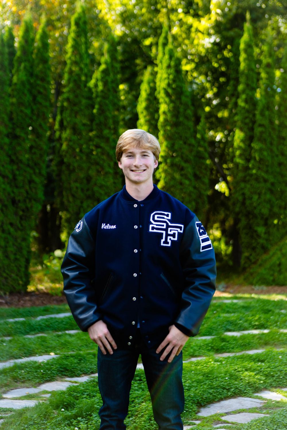 Student wearing a navy and white varsity letterman jacket poses outdoors against green hedges.