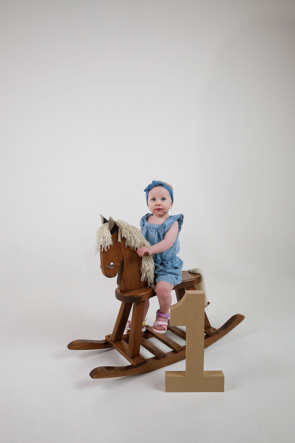 Small child in blue outfit riding wooden rocking horse against white backdrop with golden number one decoration.