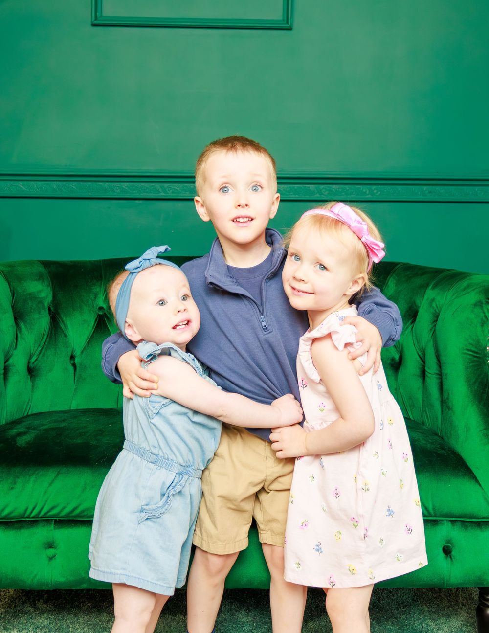 Three young children embracing on emerald green velvet sofa.