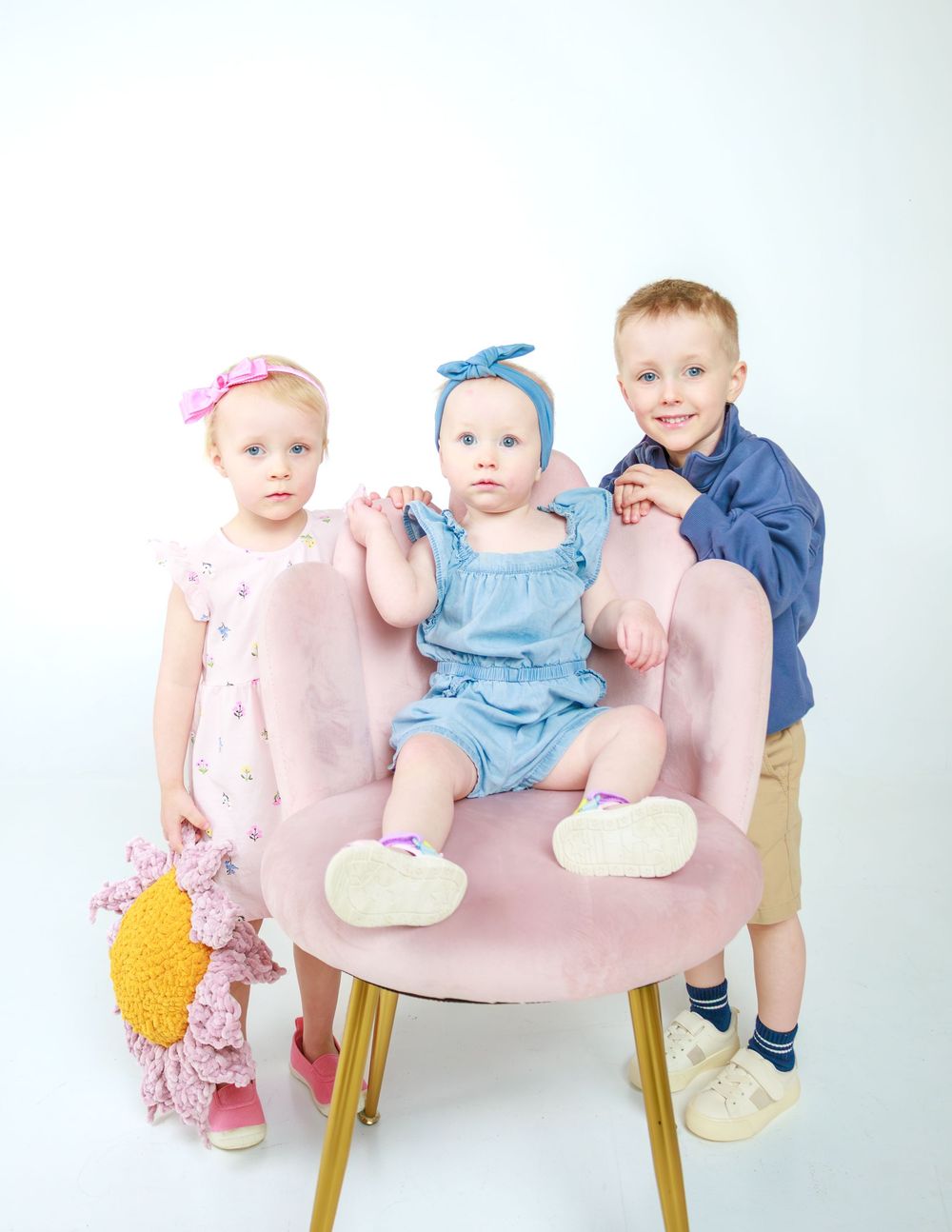 Three children posed together on pink chair against white background.