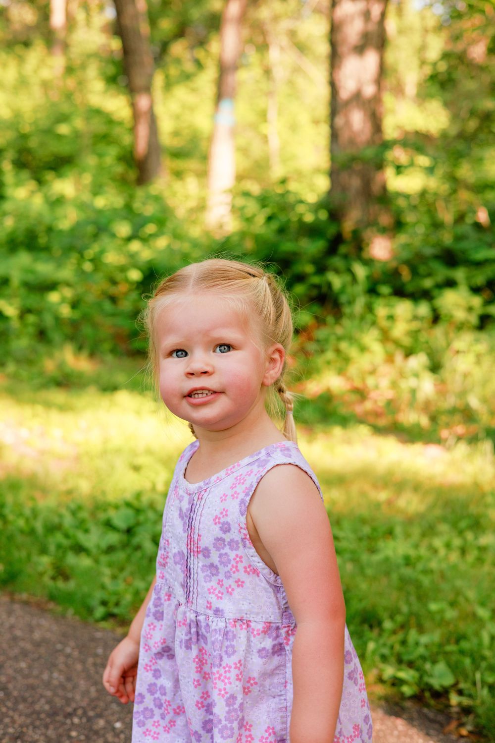 Young child in floral dress smiles at camera in sunlit wooded setting.