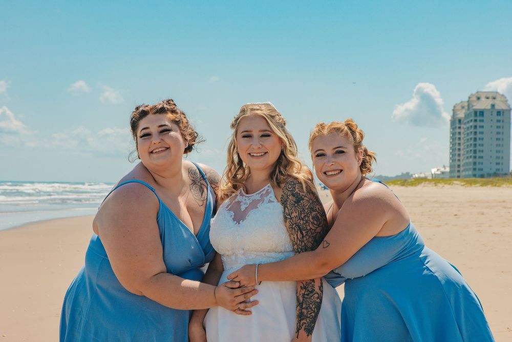 Three women in blue and white dresses embracing on sunny beach.