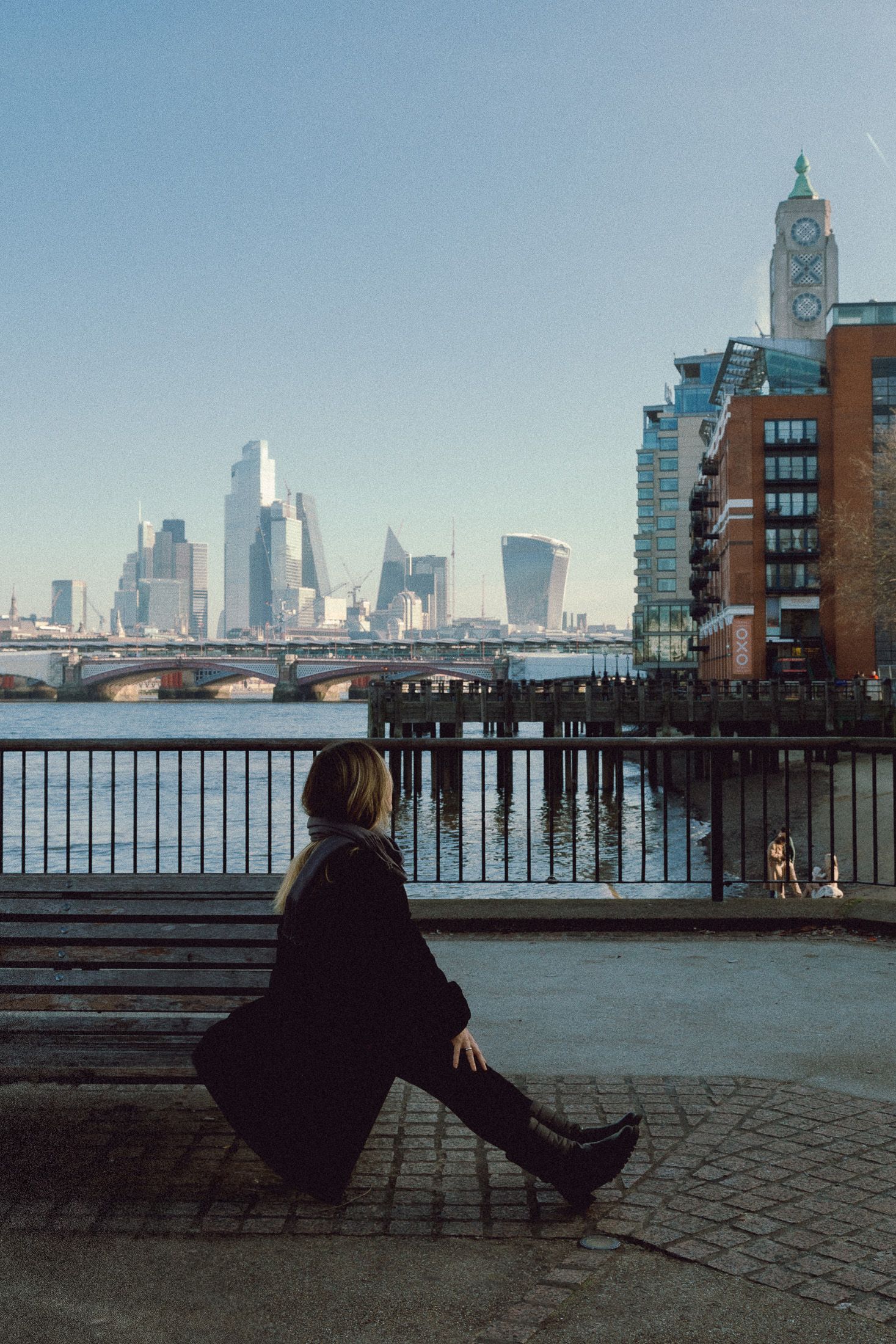 Person sits on a bench overlooking the modern London city skyline with Thames River and skyscrapers in background.