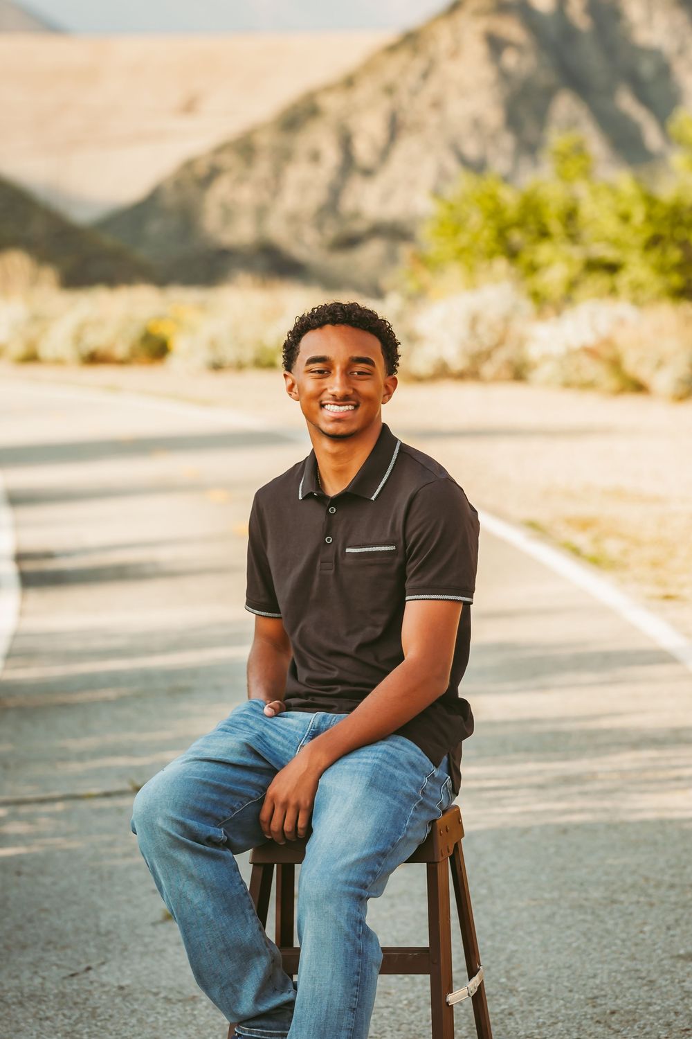 A person in a dark polo shirt and jeans sits on a stool outdoors with mountains and sunlit scenery in the background.