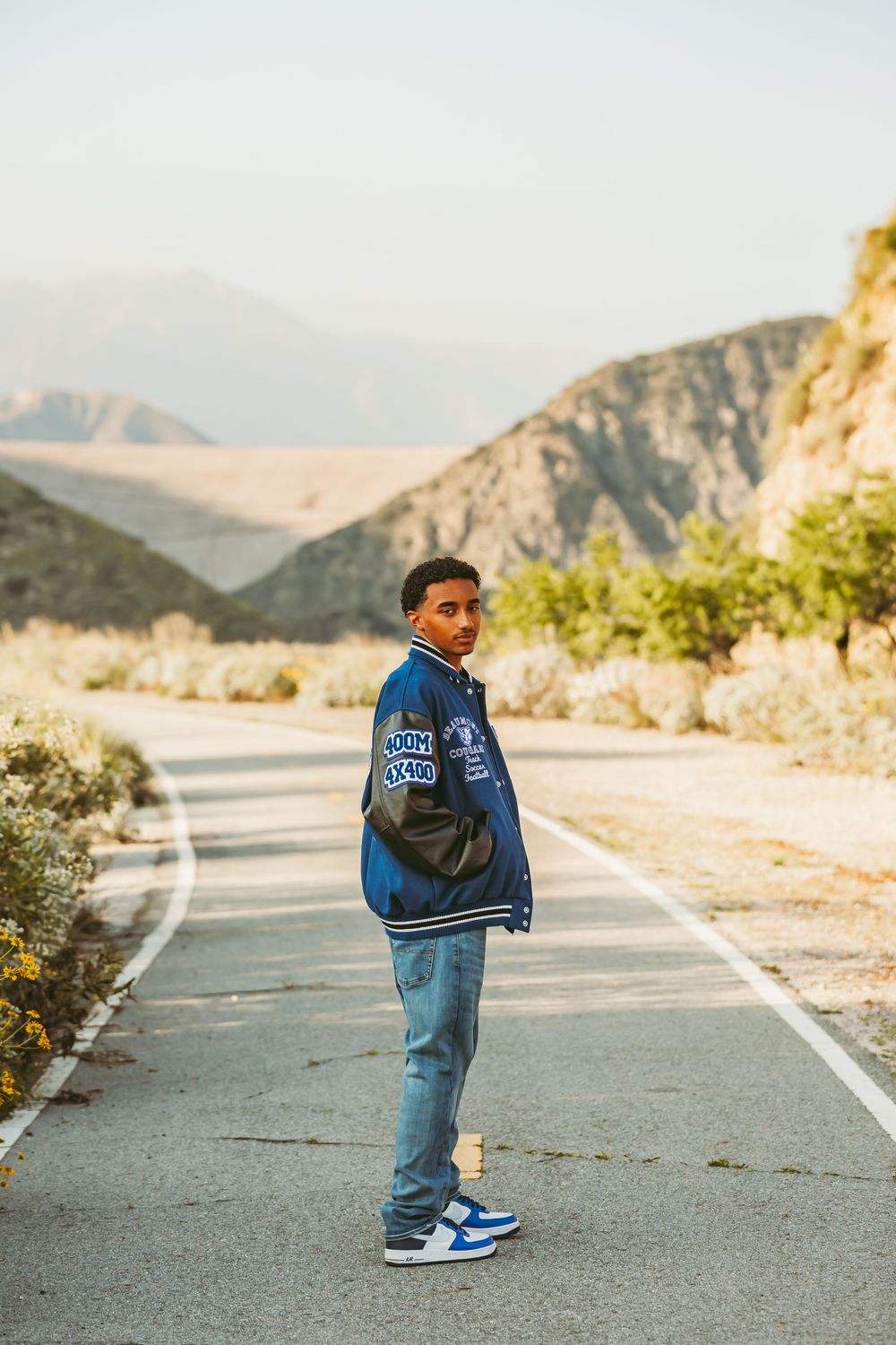 Person wearing blue varsity jacket and jeans stands on mountain path during golden hour.