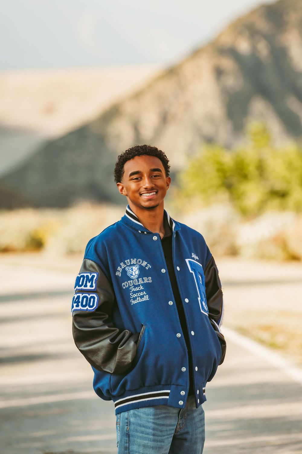 Someone wearing a blue and black varsity letterman jacket smiles while standing outdoors with mountains in the background.