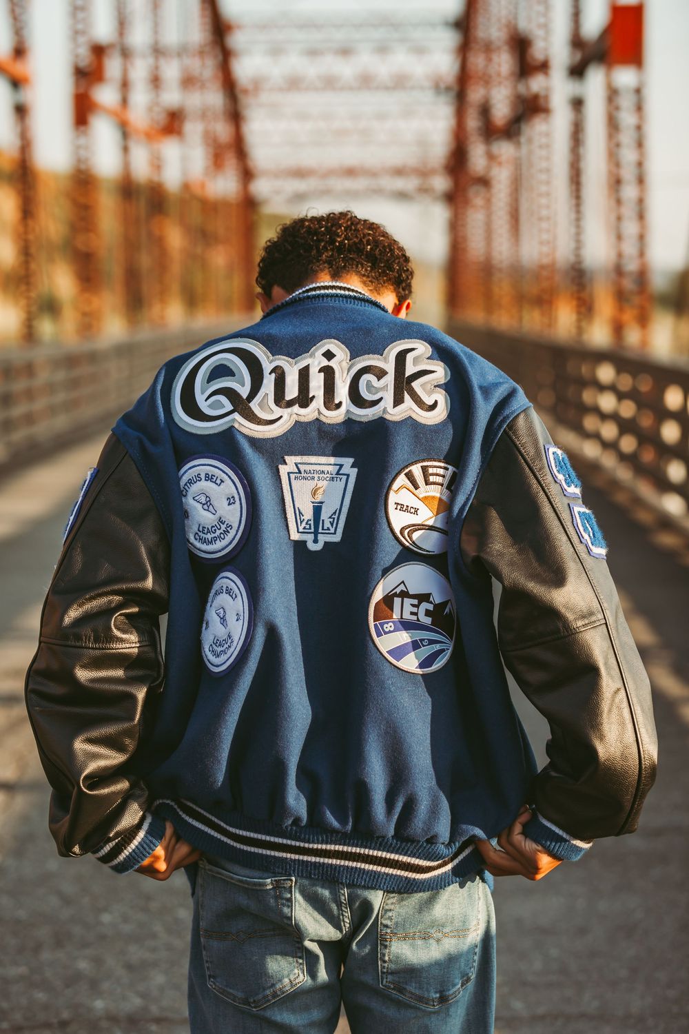 Back view of varsity jacket with 'Quick' text and sports patches on a bridge during golden hour sunset.