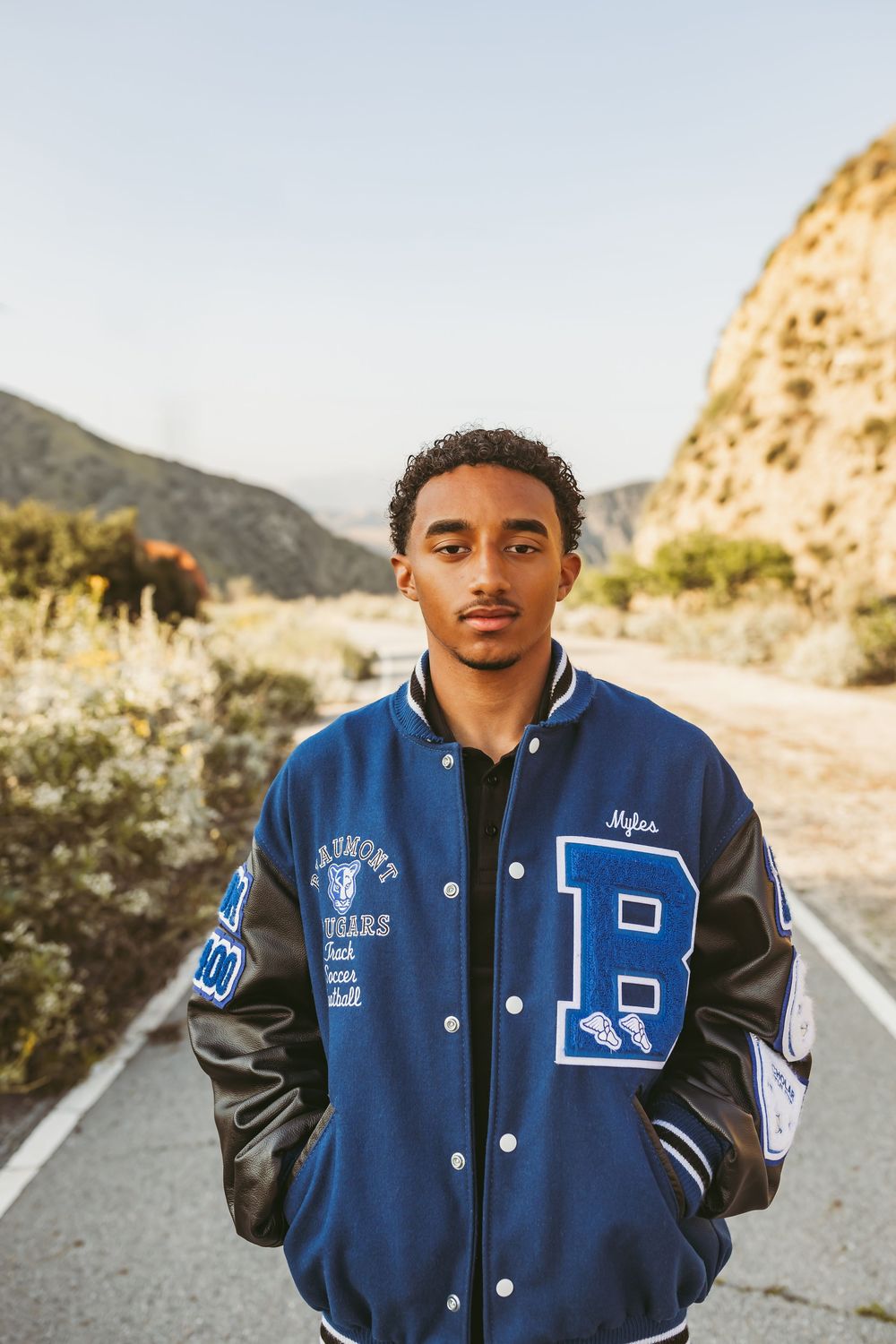 A person wearing a blue and gray varsity letterman jacket poses on a mountain road at sunset.