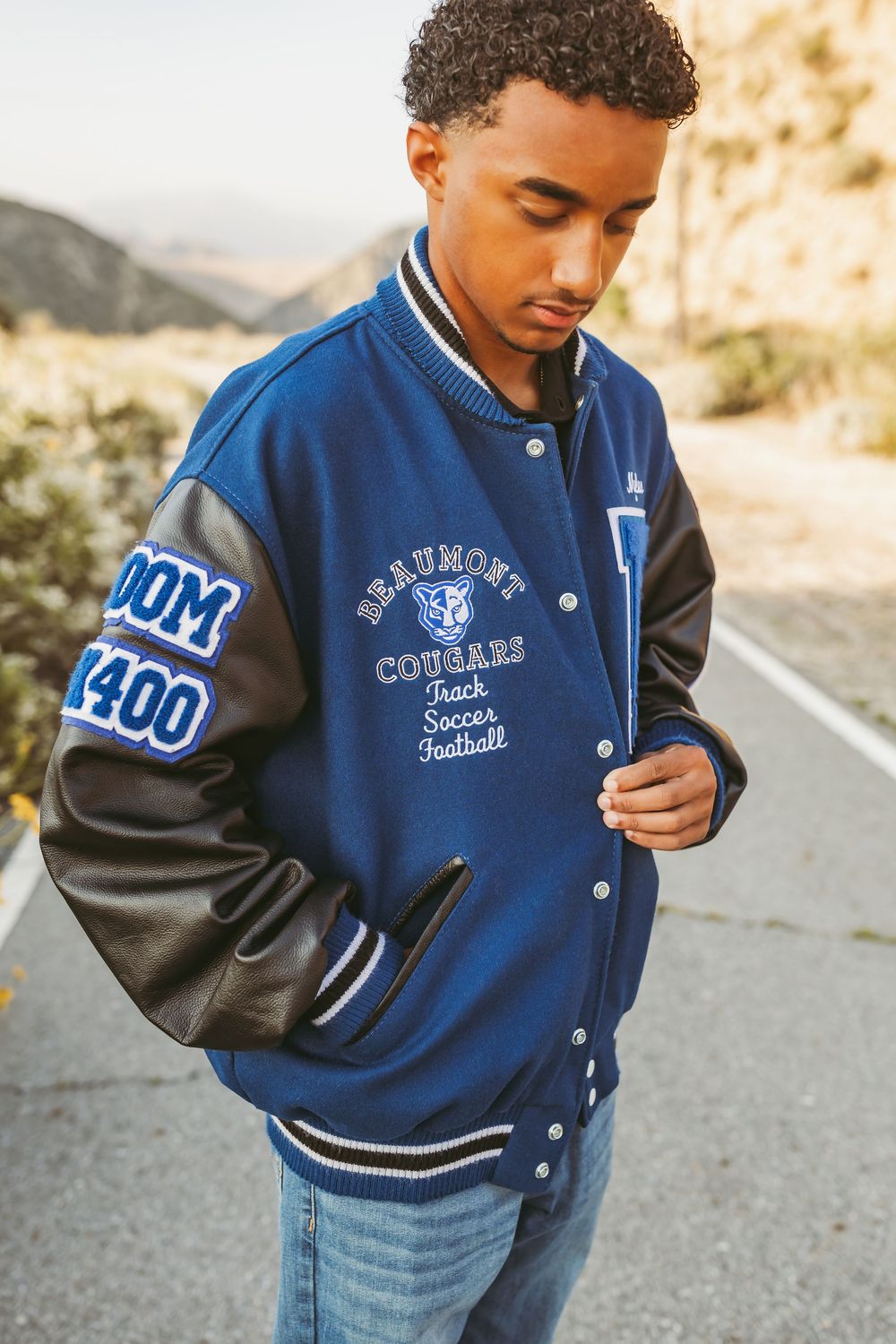 Person wearing a blue and black varsity letterman jacket with embroidered patches and jeans standing on a mountain road.