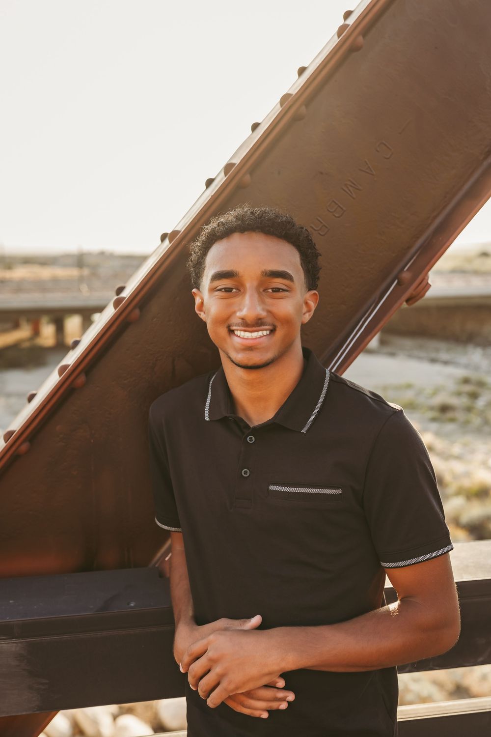 Young man in black polo shirt smiling during golden hour outdoors next to industrial architectural structure.