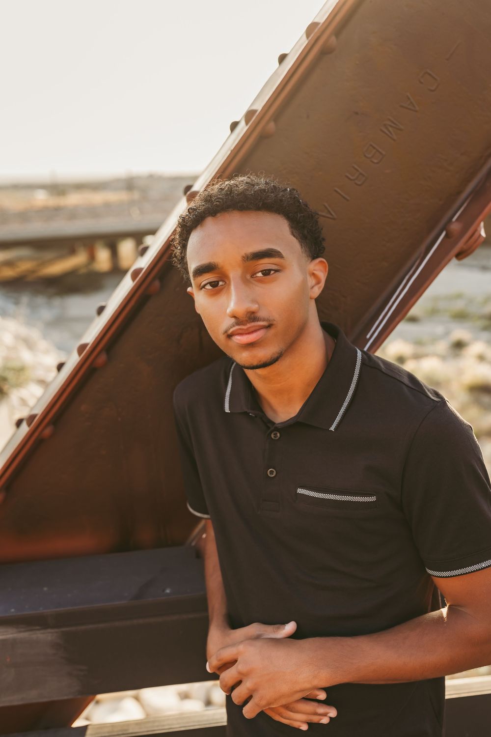 Young man in black polo shirt leaning against rustic metal structure during golden hour outdoors.