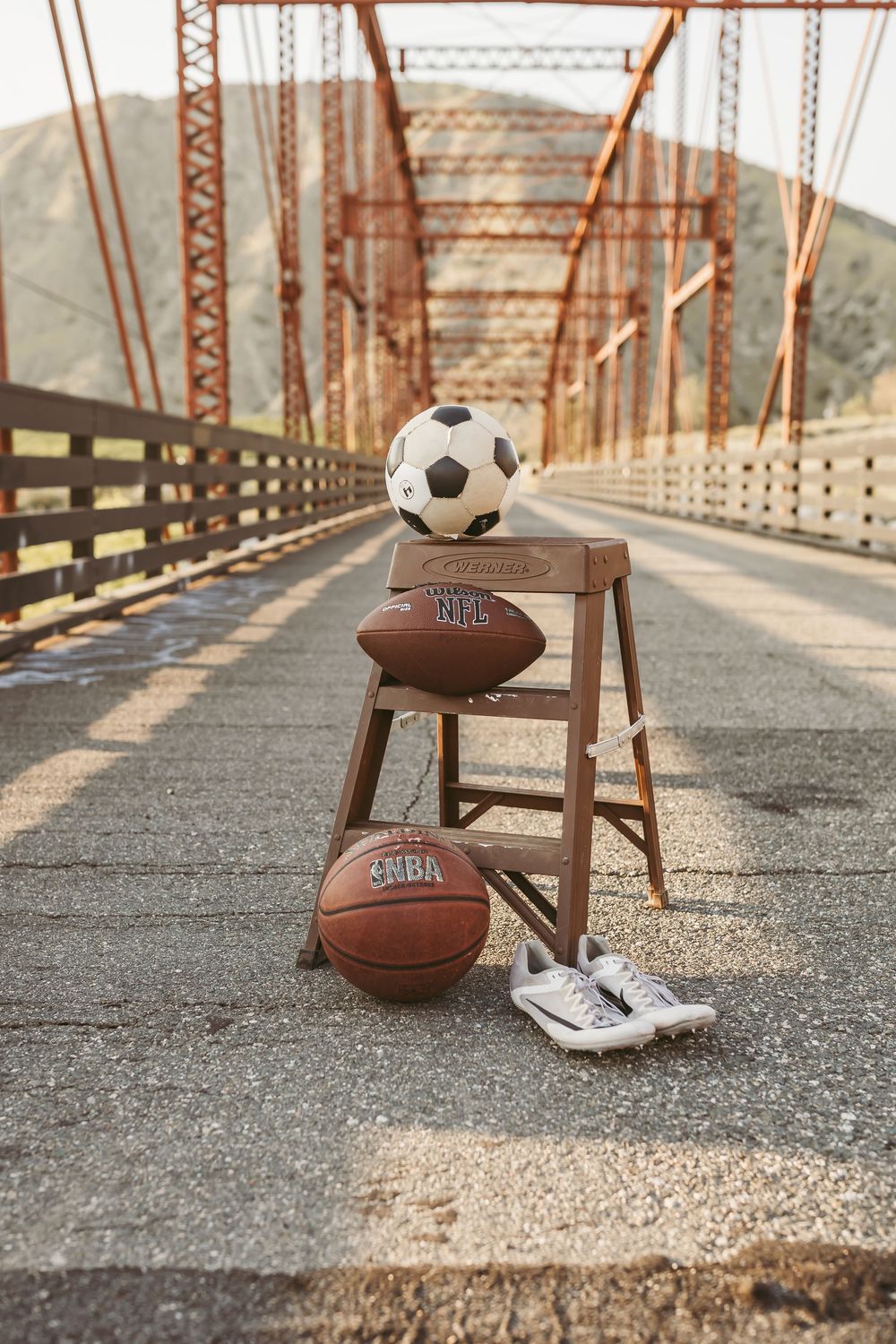 Sports equipment including soccer ball, football, and basketball arranged on wooden stool on bridge walkway at sunset.