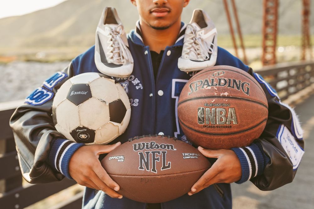 Person in varsity jacket holds multiple sports equipment including football, soccer ball, basketball and sneakers.