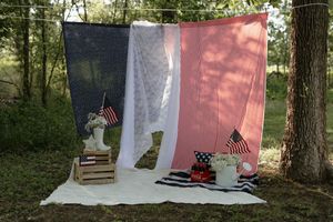 Patriotic red white and blue blankets hung outdoors with rustic wooden chair and vintage decor in a woodland setting.