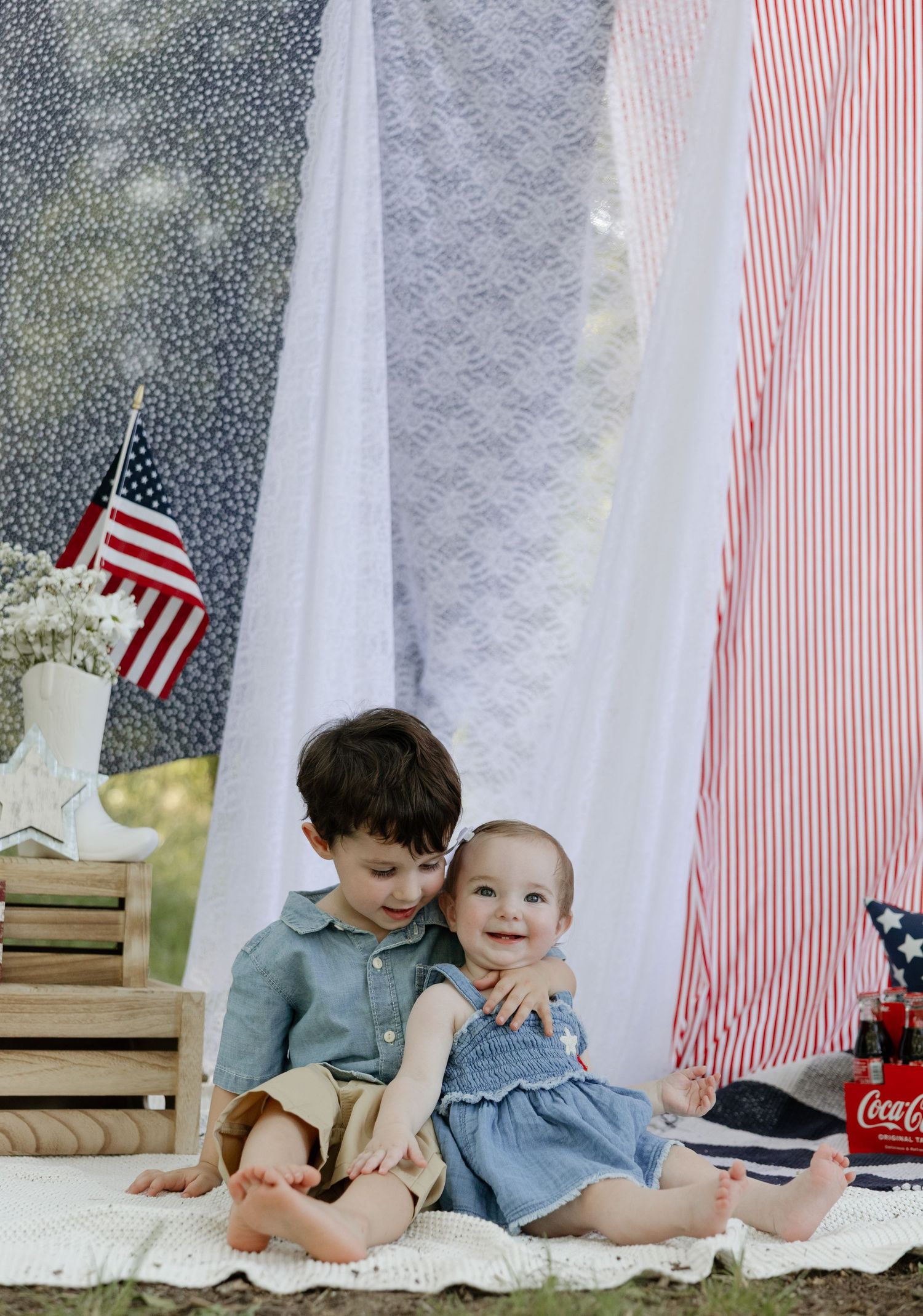 Two children sit together on a blanket with American flag decor, Coca-Cola bottle and striped fabric backdrop for patriotic photo.