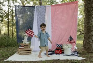 Patriotic outdoor photo scene with American flags, wooden crates and red white blue fabric backdrop against trees.