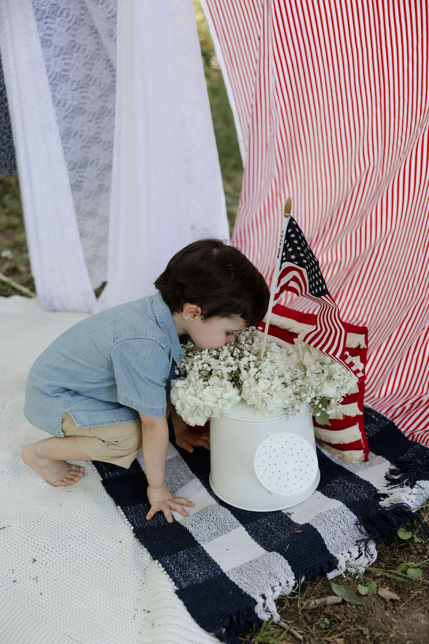A young child in blue shirt arranges flowers in a white vase next to red and white striped fabric and American flags.