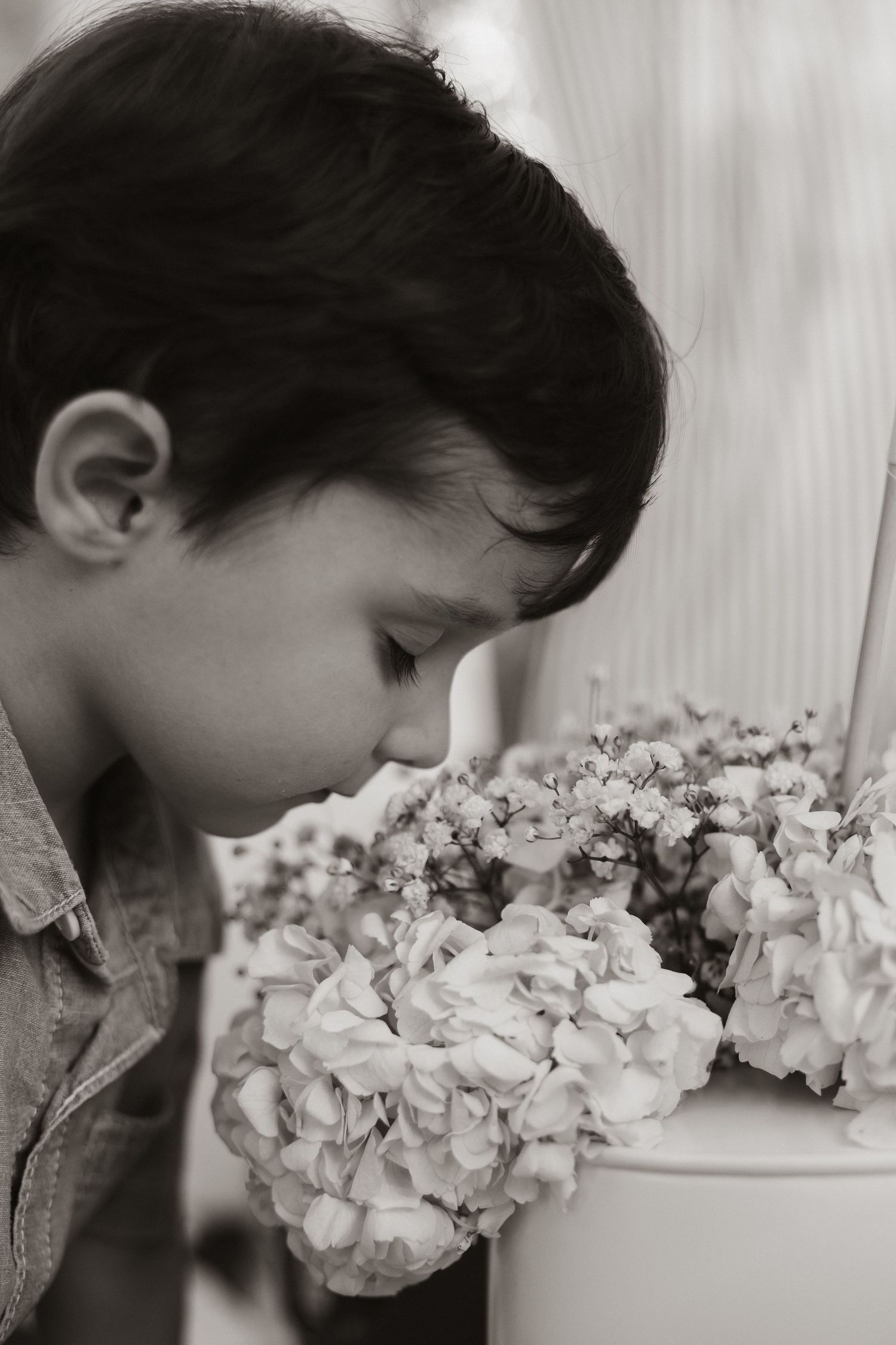 A young boy in profile leaning in to smell white hydrangea flowers in a black and white photograph.