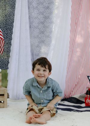 A smiling young child in chambray shirt and khaki shorts sits cross-legged on a blanket with decorative curtains and a flag behind.