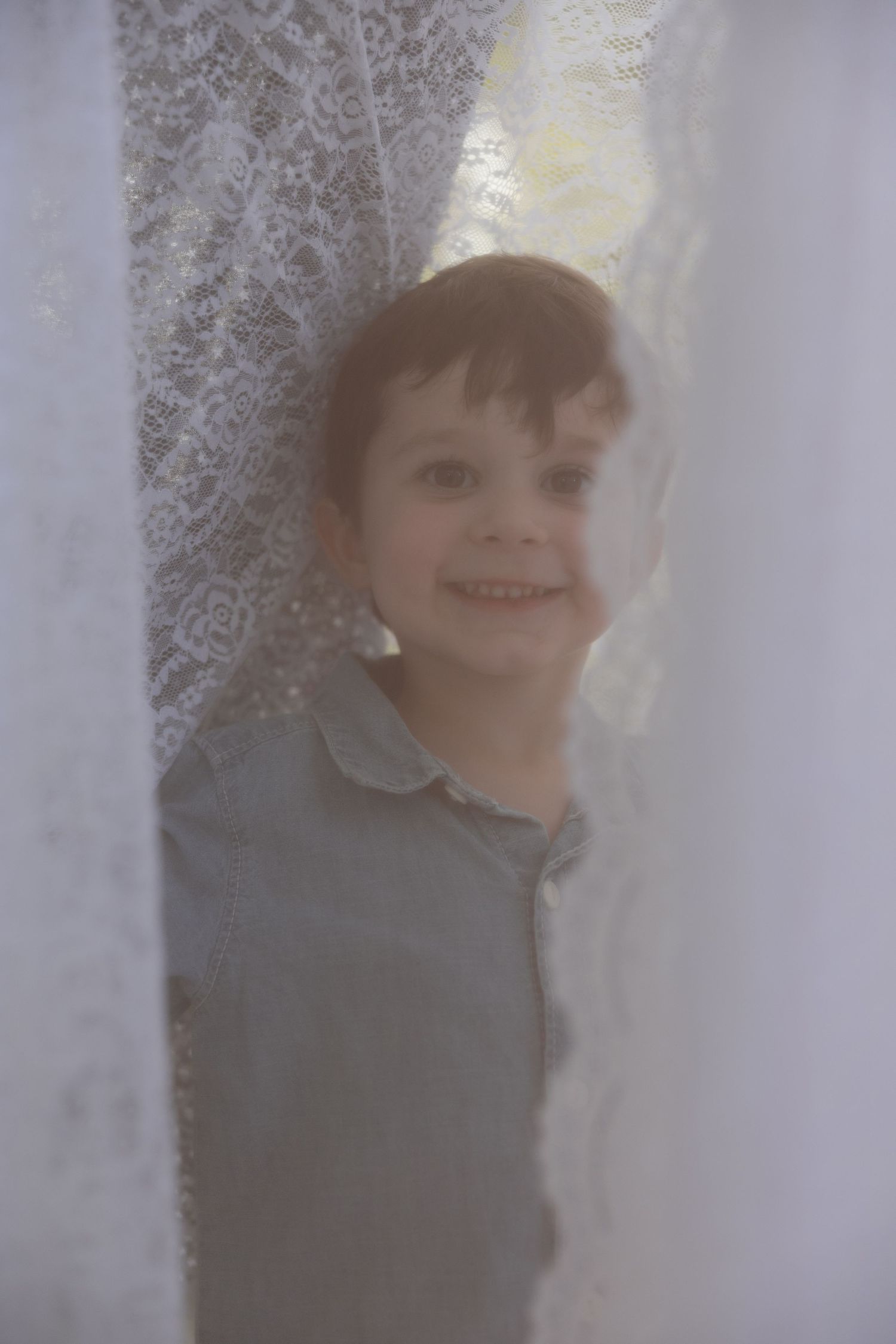 A child peeks through white lace curtains in a soft, ethereal portrait with a dreamy quality.