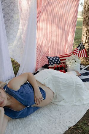 A summer picnic scene with a striped backdrop, American flags, and someone reclining on a white blanket outdoors.