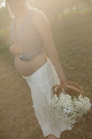 A dreamy golden-hour maternity portrait shows a woman in white dress holding a basket of baby's breath flowers in a field.