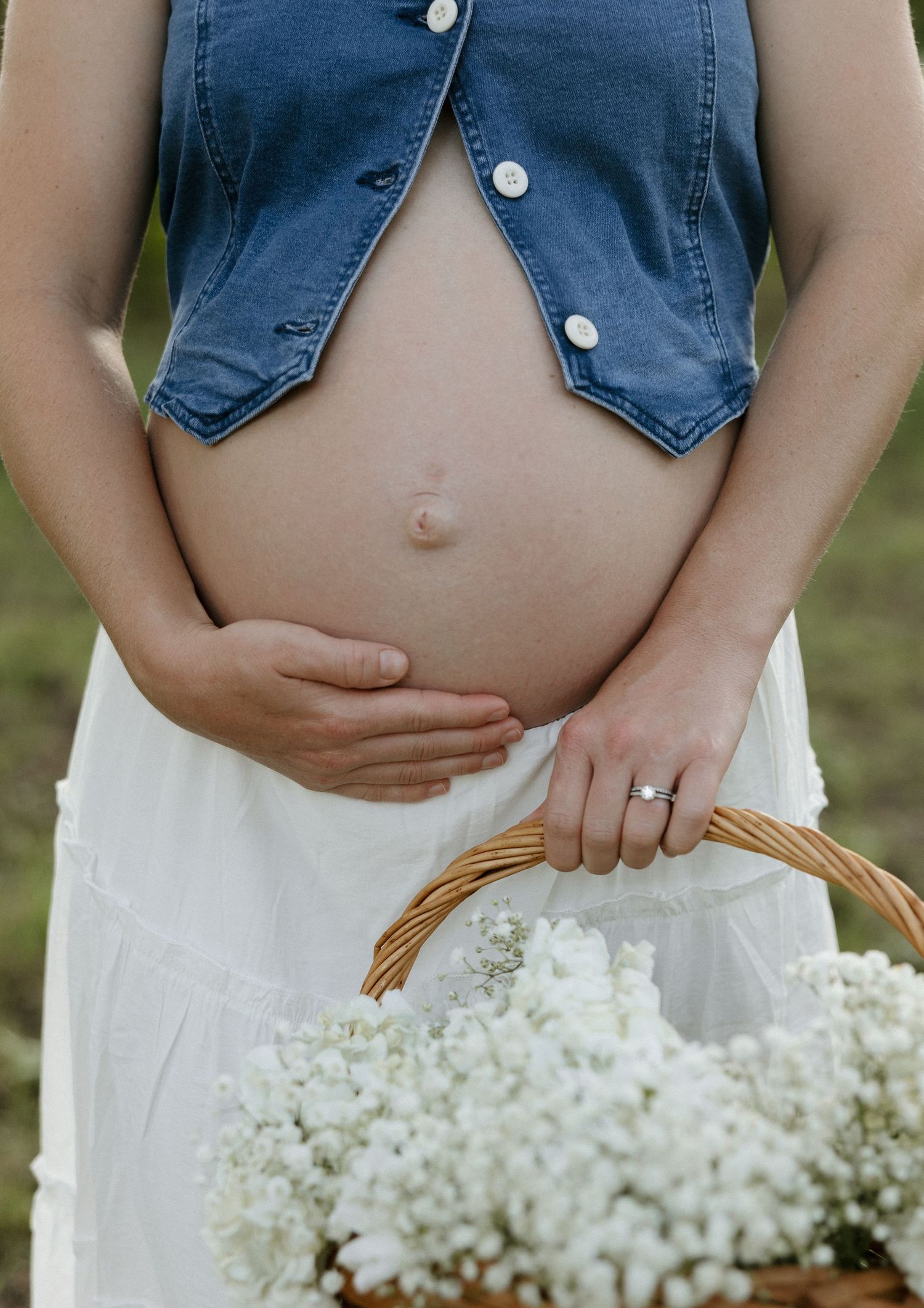 A pregnant belly shown with an open denim vest and white dress, holding a basket of delicate white baby's breath flowers.