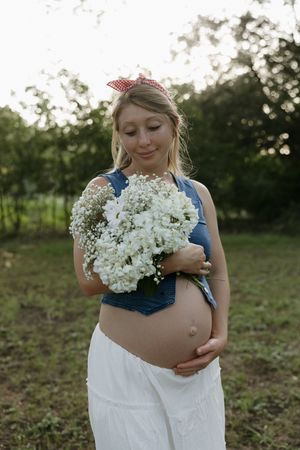 Expecting parent in blue top holds delicate white baby's breath flowers while standing outdoors at sunset.