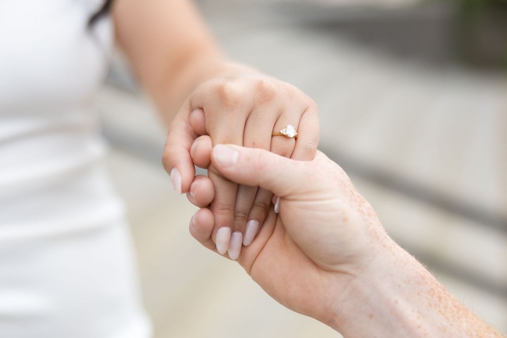 Two hands gently clasped together wearing engagement rings against a soft blurred background.