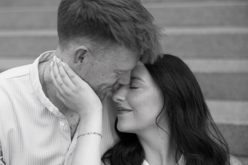 A loving couple shares an intimate candid moment leaning in close together on stone steps in black and white.