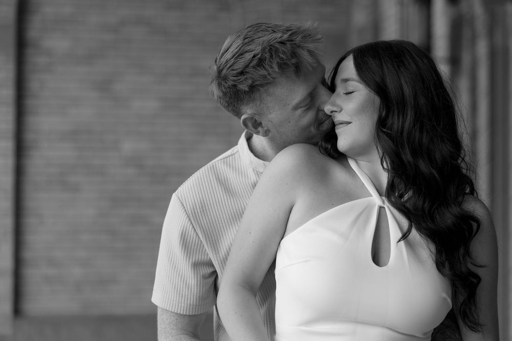 A tender romantic black and white portrait of a couple in white clothing sharing an intimate moment against a brick wall.