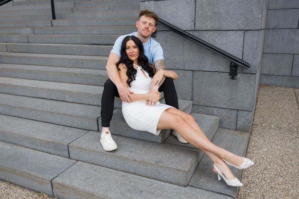 A couple in formal attire sits intimately on gray stone steps in a romantic engagement photoshoot.