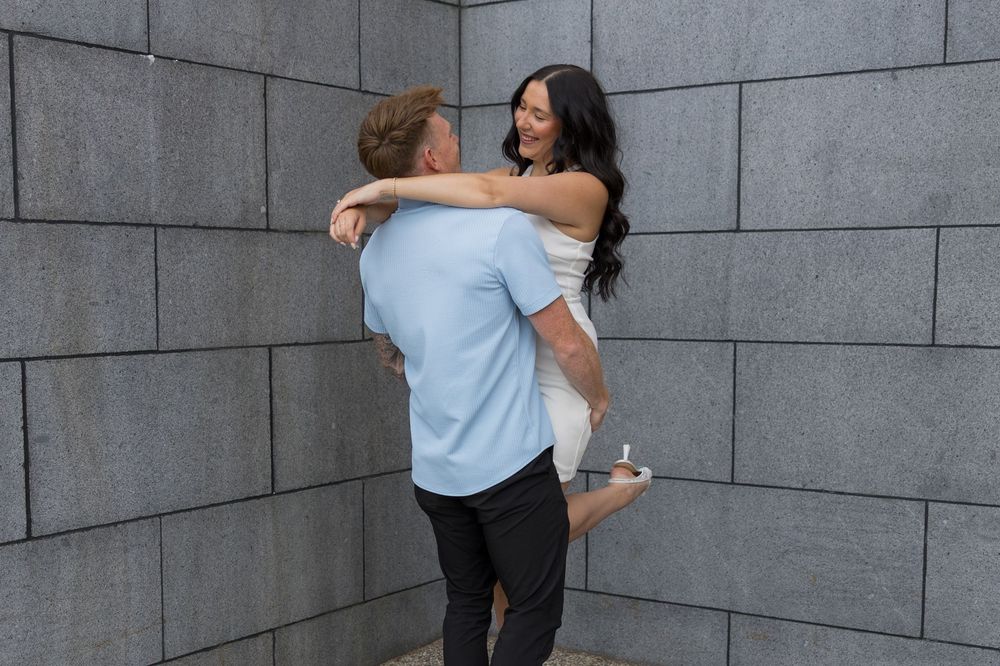 Couple shares intimate moment against grey concrete wall as she jumps into his arms for playful embrace.