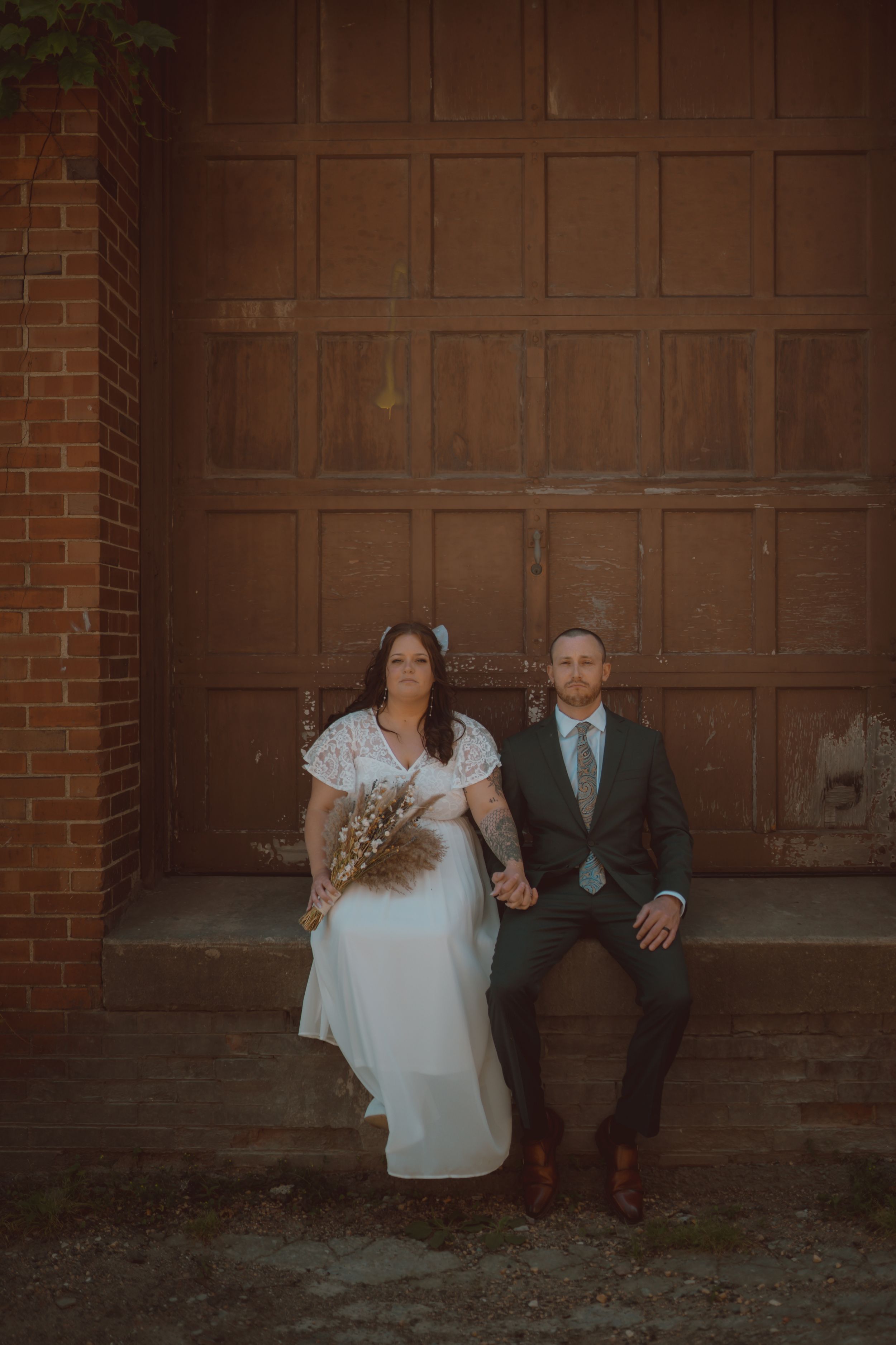 Couple in wedding attire sitting against a brown wooden garage door in a romantic outdoor setting.