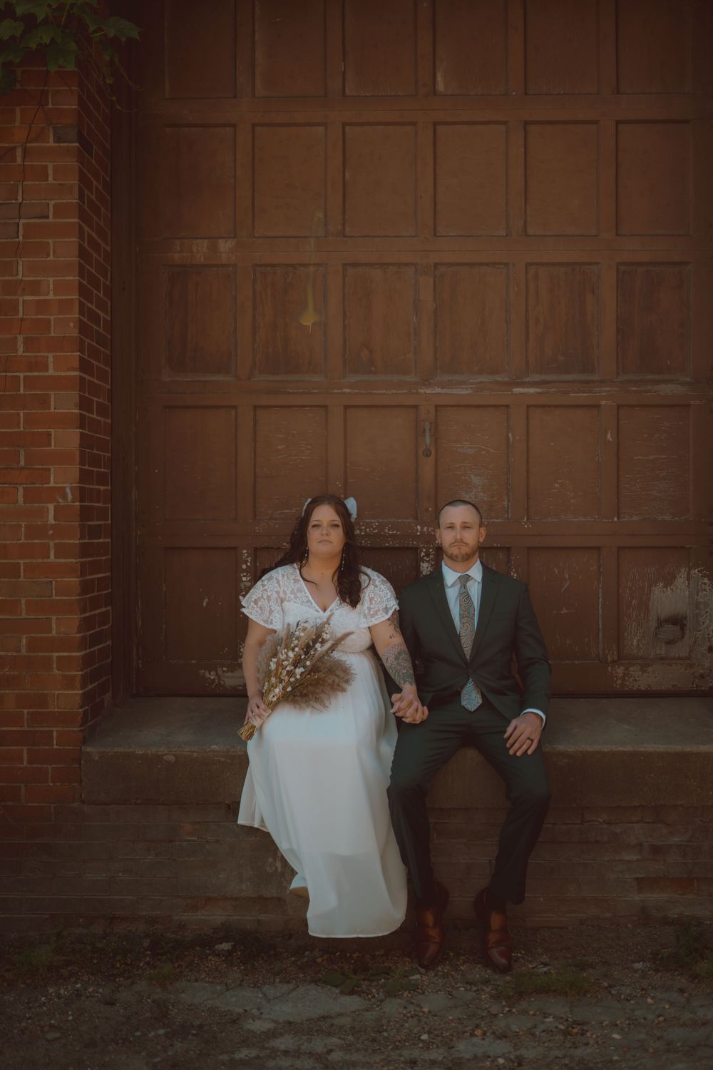 Couple in wedding attire sitting against a brown wooden garage door in a romantic outdoor setting.