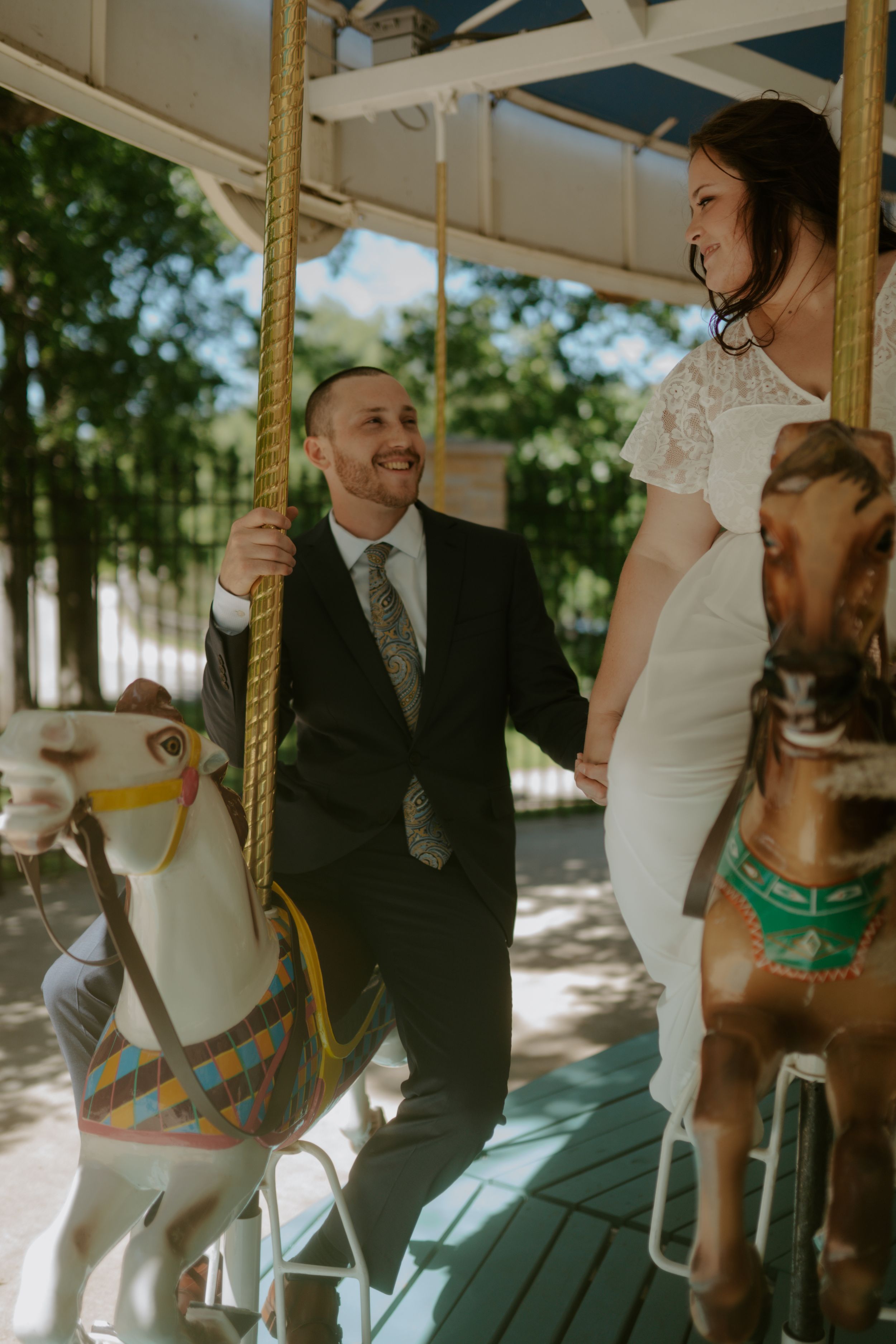 Playful wedding couple enjoying moments on a vintage carousel in a sunny outdoor setting.