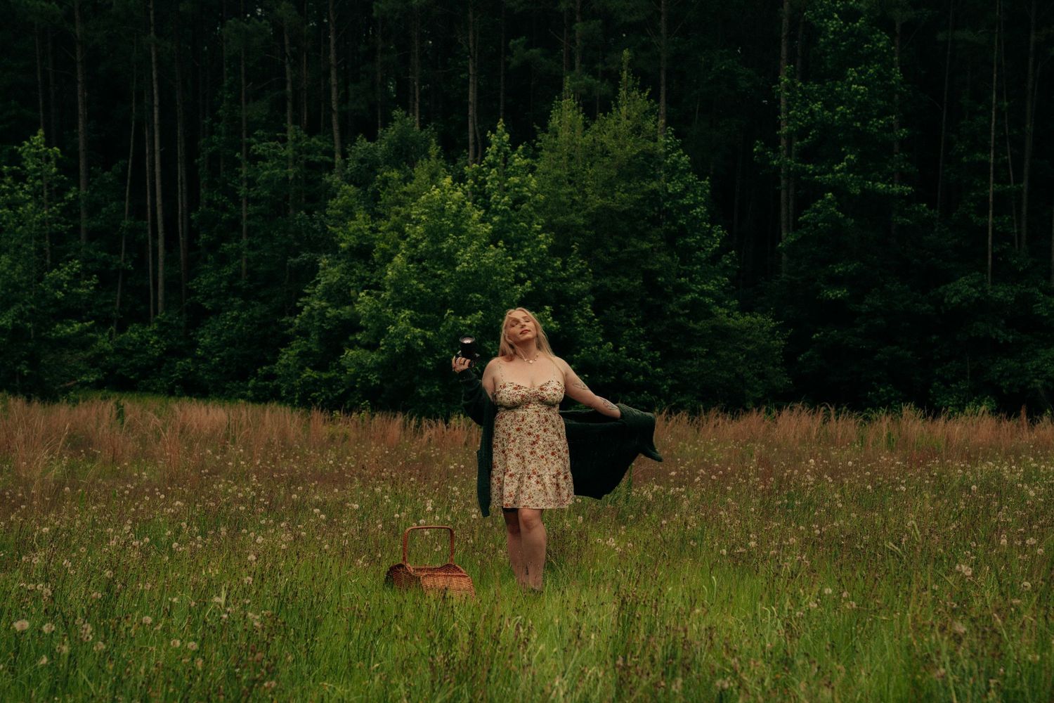 Figure in a floral dress stands in a meadow with forest backdrop while holding a black umbrella.