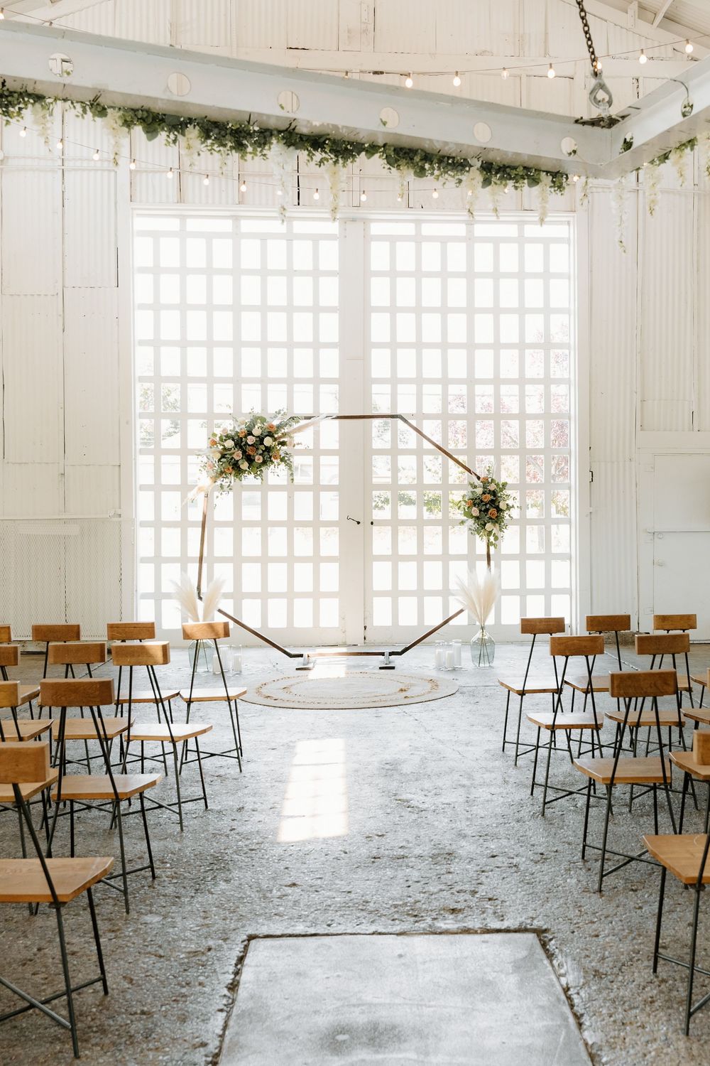 Wedding ceremony setup with hexagonal arch and wooden chairs in a bright industrial space.