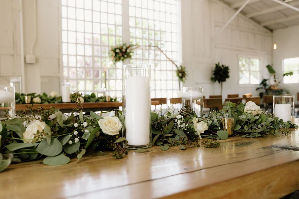 Long wooden dining table decorated with elegant white candles, eucalyptus garlands, and white floral centerpieces in a bright venue.