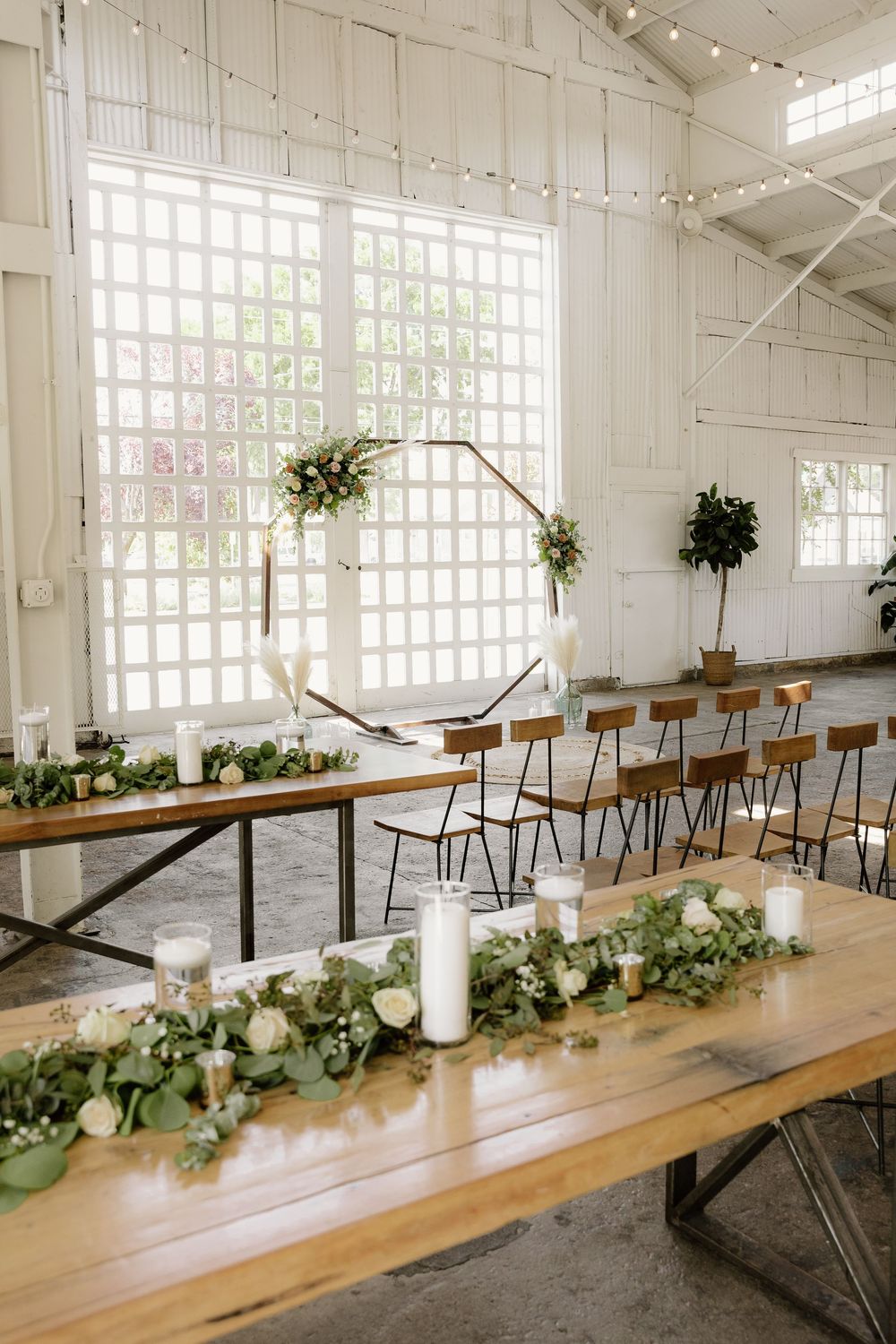 Wedding reception table decorated with greenery and candles in front of geometric backdrop.