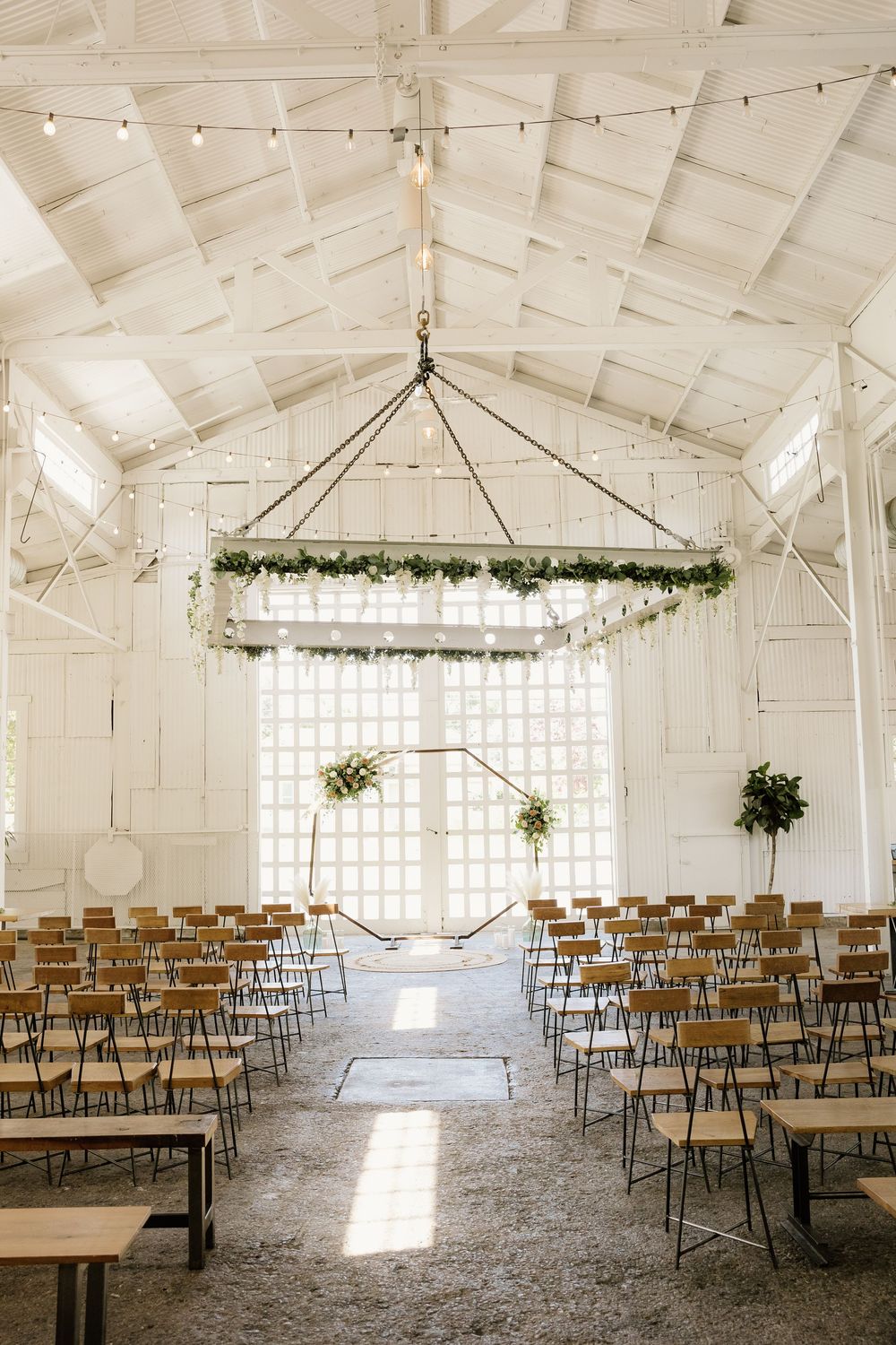 Bright white wedding ceremony venue with wooden chairs and hanging greenery chandelier.