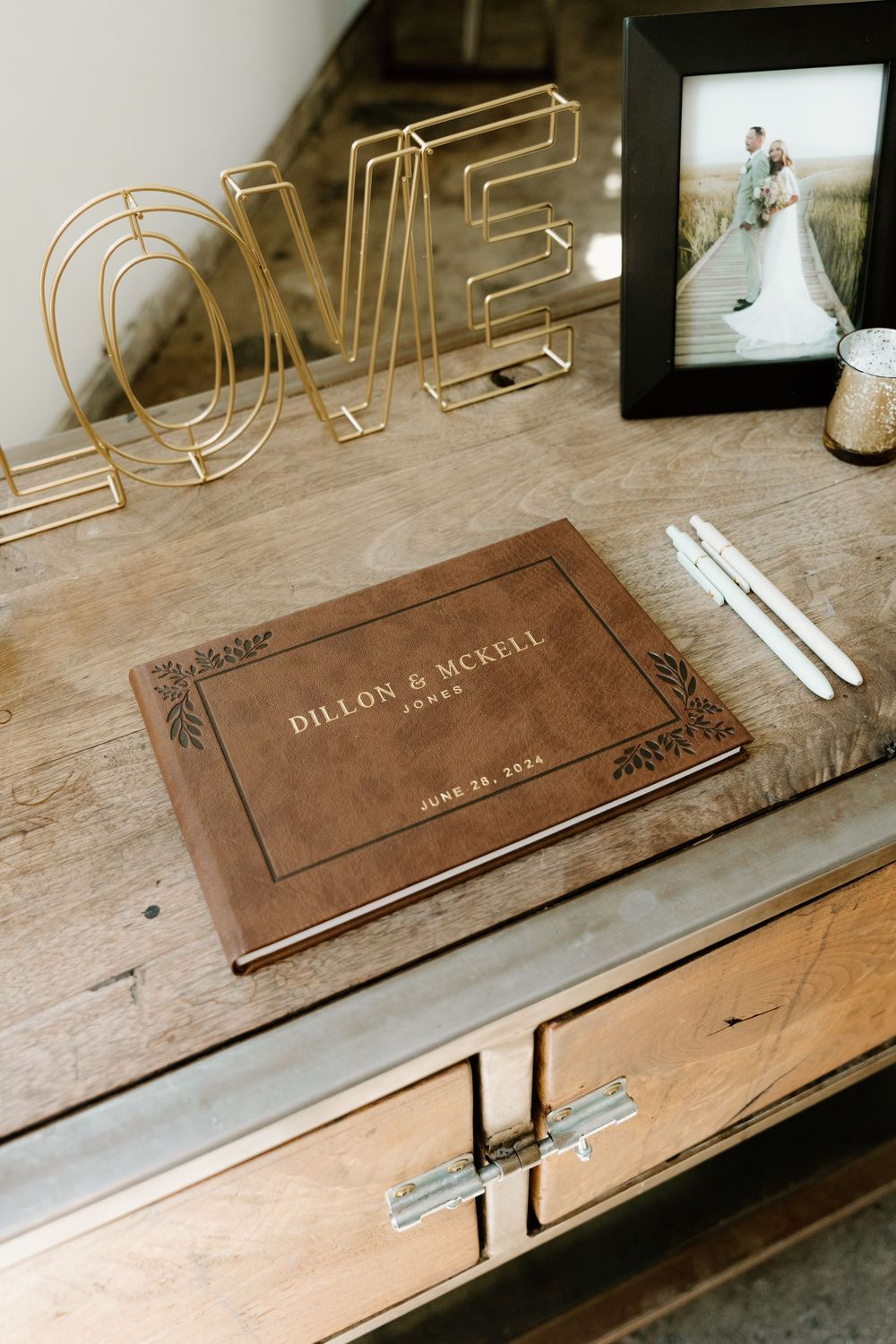 A wedding guestbook and gold 'LOVE' sign on a wooden table with a framed photo.